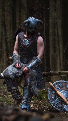 A guy worn a Viking inspired clothing sits on a tree branch in the woods, with a Norse themed shield and sword by his side 